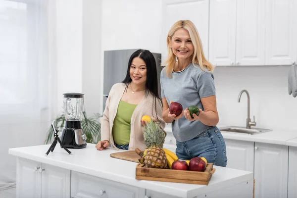 Alegre bloggers multiétnicos sosteniendo frutas frescas y mirando a la cámara cerca de teléfonos inteligentes y licuadora en la cocina - foto de stock