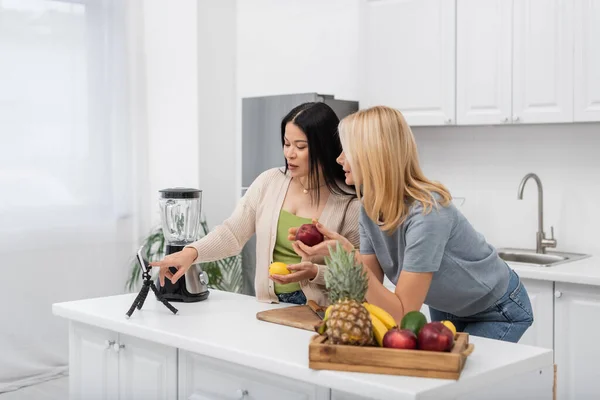 Bloggers multiétnicos usando teléfonos inteligentes y sosteniendo frutas cerca de la licuadora en casa - foto de stock