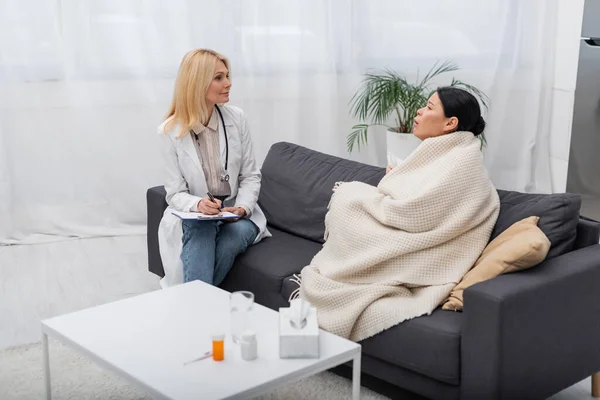 Doctor with clipboard looking at sick asian patient at home — Stock Photo