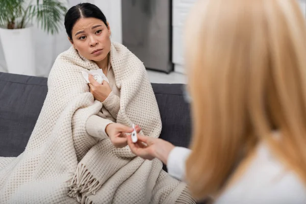Enfermo asiático mujer con servilleta dando termómetro a borrosa médico en casa - foto de stock