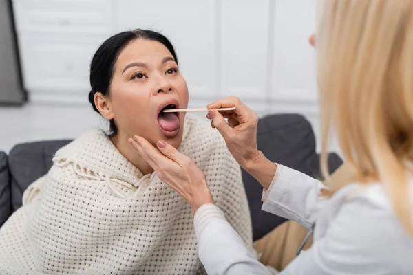 Blurred doctor en blanco capa chequeo garganta de asiático paciente en casa - foto de stock