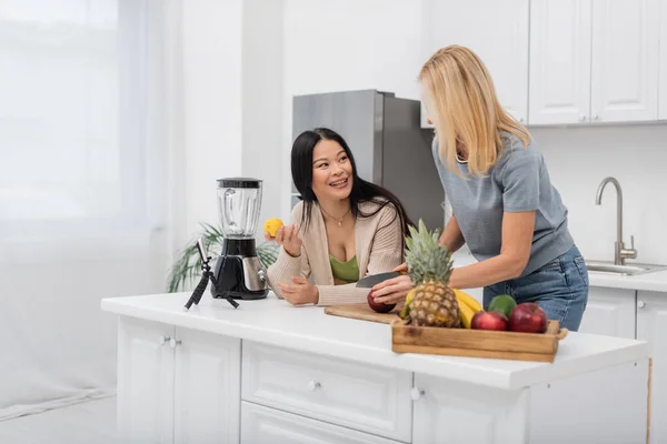 Sourire asiatique femme tenant citron et parler à ami coupe pomme près smartphone sur trépied dans cuisine — Photo de stock