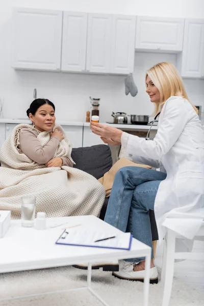 Smiling doctor holding pills near sick asian woman in blanket at home — Stock Photo
