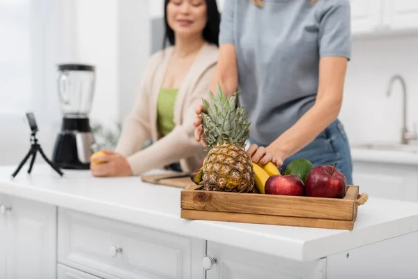 Cropped view of blurred woman taking fruits near blurred asian friend and smartphone on tripod at home — Stock Photo
