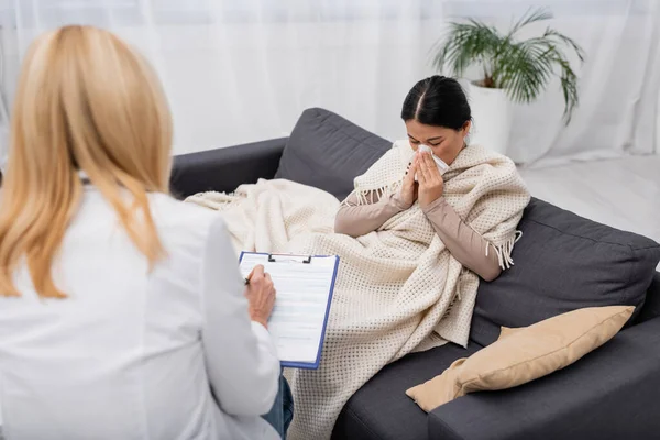 Sick asian woman suffering from runny nose near blurred doctor with clipboard at home — Stock Photo
