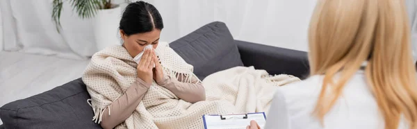 Ill asian woman sneezing near blurred doctor with clipboard at home, banner — Stock Photo