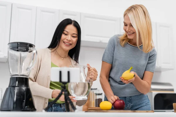 Smiling interracial bloggers holding water and banana near smartphone on tripod at home — Stock Photo