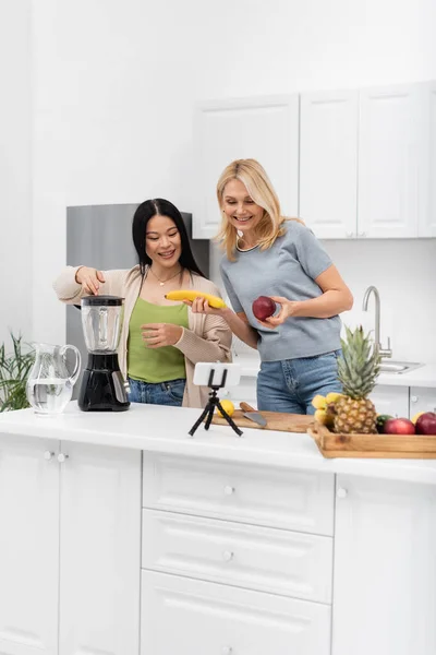 Positive interracial bloggers holding fruits near blender and smartphone on tripod at home — Stock Photo