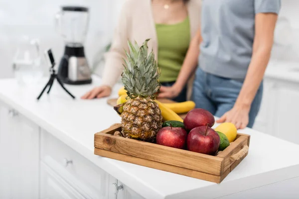 Cropped view of fresh fruits near blurred friends and smartphone on tripod in kitchen — Stock Photo