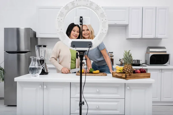 Positive interracial bloggers looking at smartphone in ring lamp near fruits in kitchen — Stock Photo