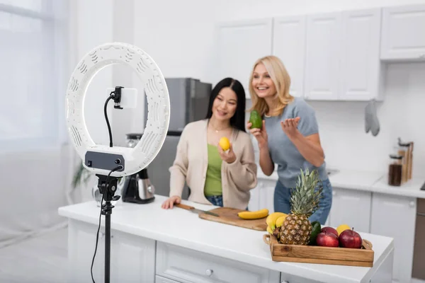 Ring lamp and smartphone near blurred interracial bloggers with ripe fruits at home — Stock Photo