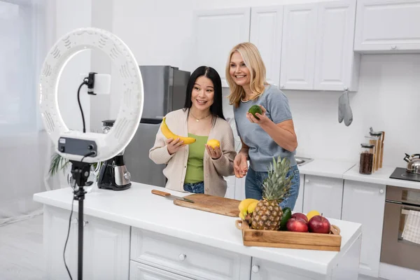 Multiethnic bloggers holding organic fruits near smartphone and ring lamp at home — Stock Photo