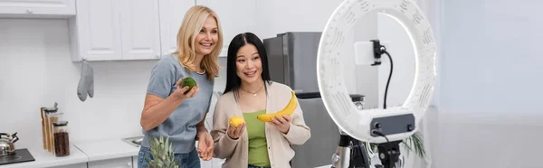 Interracial bloggers holding fresh fruits near cellphone in ring lamp at home, banner — Stock Photo