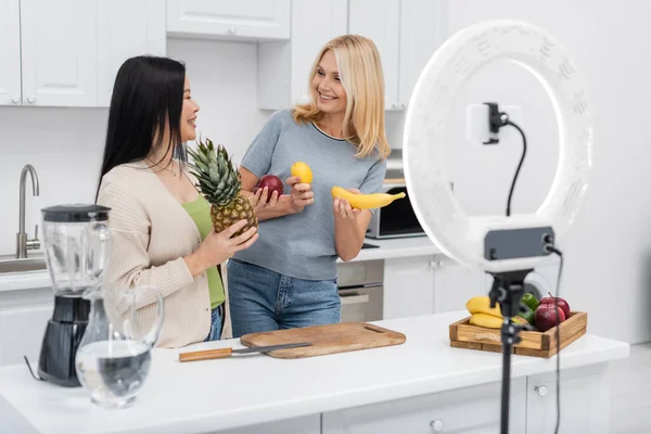 Interracial bloggers smiling and holding fruits near smartphone in ring lamp in kitchen — Stock Photo