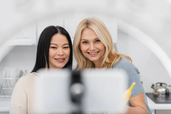 Smiling multiethnic bloggers holding banana and looking at blurred smartphone with ring lamp — Stock Photo
