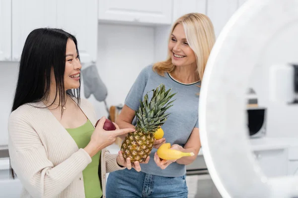 Cheerful asian woman pointing at friend with fresh fruits near blurred cellphone and ring lamp in kitchen — Stock Photo