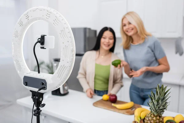 Celular y anillo lámpara cerca borrosa interracial amigos con frutas en la cocina - foto de stock