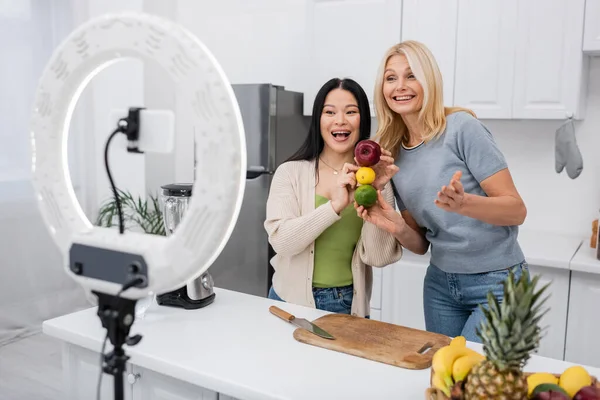 Excited interracial bloggers holding ripe fruits near smartphone in kitchen — Stock Photo