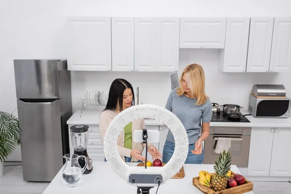 Multiethnic friends looking at fruits near smartphone and ring lamp in kitchen — Stock Photo