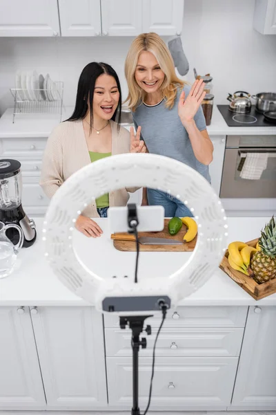 Positivo interrazziale blogger gesticolando smartphone in lampada ad anello vicino frutta fresca in cucina — Foto stock