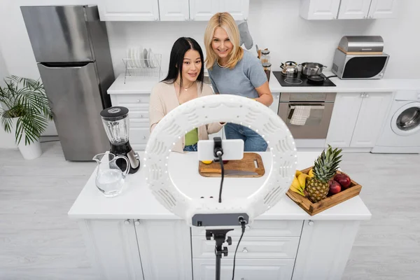 Interracial friends looking at smartphone in ring lamp near blender and fruits in kitchen — Stock Photo