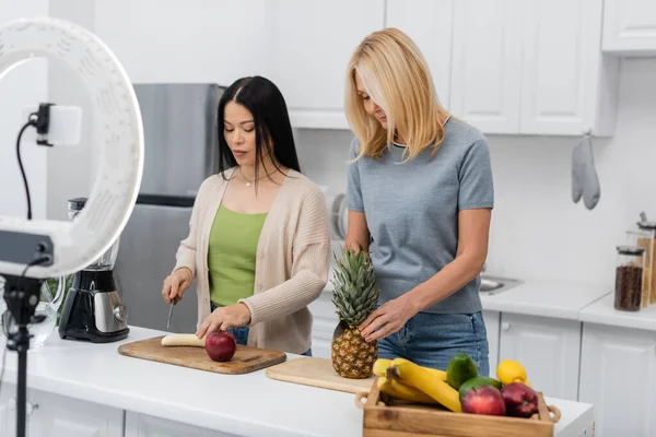 Interracial friends cutting fruits near smartphone in ring lamp in kitchen — Stock Photo