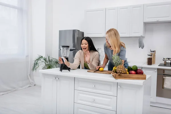 Cheerful asian blogger waving hand at smartphone near friend and fresh fruits in kitchen — Stock Photo
