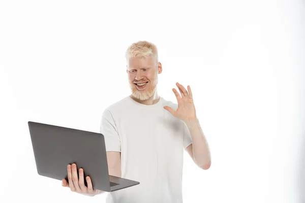 Barbuto albino uomo in t-shirt sorridente e mano ondulata durante la videochiamata su computer portatile su sfondo bianco — Foto stock