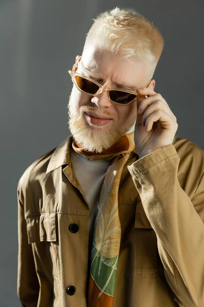 Sunlight on face of pleased albino man adjusting stylish sunglasses on grey — Stock Photo