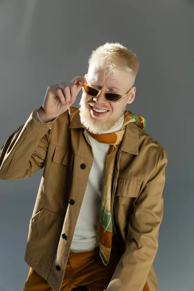 Sunlight on face of smiling albino man adjusting stylish sunglasses on grey — Stock Photo