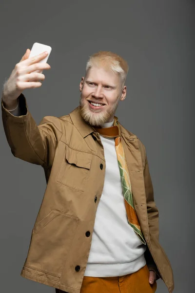 Cheerful albino man in stylish shirt jacket taking selfie on smartphone isolated on grey — Stock Photo