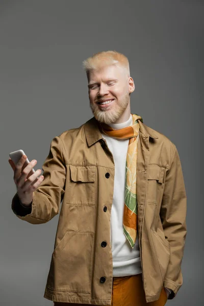 Felice albino uomo in camicia giacca tenendo smartphone isolato su grigio — Foto stock