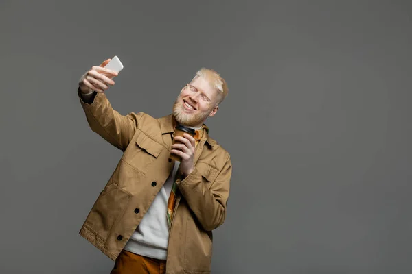 Pleased albino man in shirt jacket holding paper cup and taking selfie on smartphone isolated on grey — Stock Photo