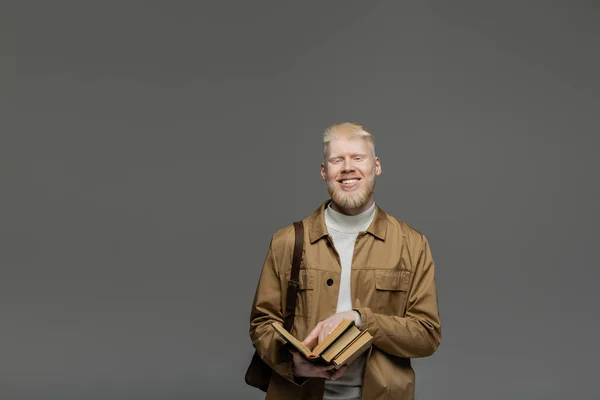 Happy albino student with backpack holding books isolated on grey — Stock Photo