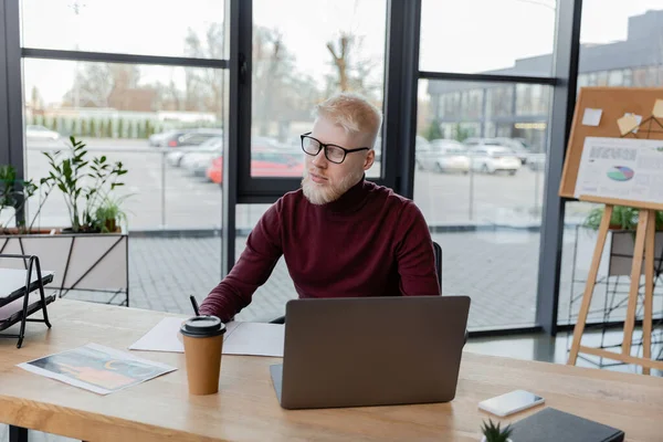 Bärtiger Albino-Geschäftsmann mit Brille in der Nähe von Gadgets und Pappbecher auf dem Schreibtisch — Stockfoto