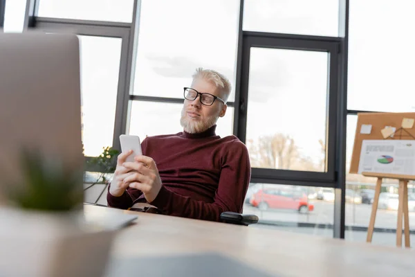 Бородатий альбіно бізнесмен в окулярах, використовуючи смартфон біля ноутбука в офісі — стокове фото