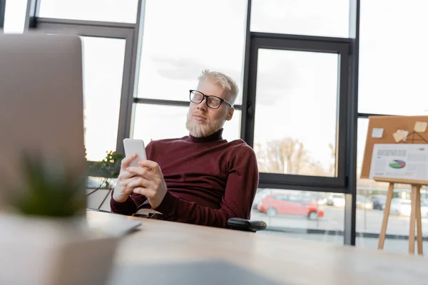 Bearded albino businessman in glasses using smartphone near laptop in office — Stock Photo