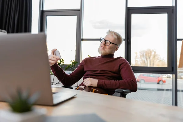 Albino businessman in glasses using smartphone and smiling near laptop in office — Stock Photo