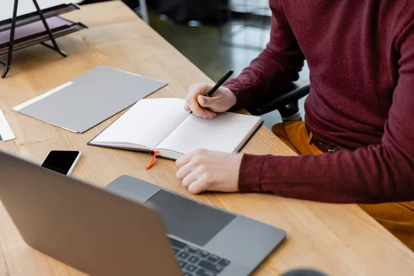 Cropped view of businessman writing on notebook near devices on desk — Stock Photo