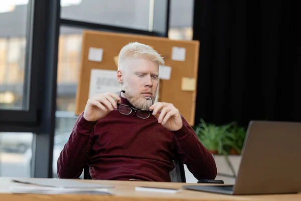 Homem de negócios albino barbudo segurando óculos enquanto olha para laptop no escritório — Fotografia de Stock