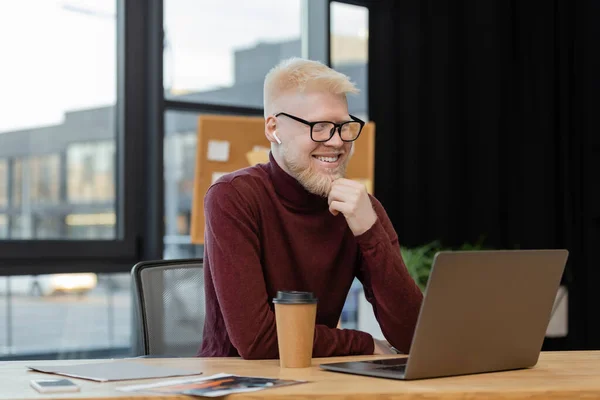 Happy albino businessman in glasses looking at laptop near paper cup on desk — Stock Photo