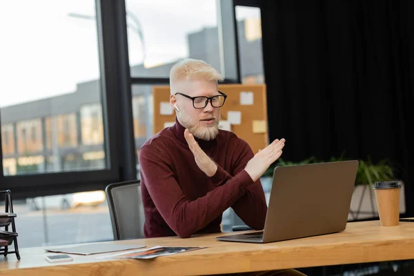 Bearded albino businessman in earphones showing refuse gesture during video call on laptop while working in office — Stock Photo
