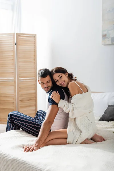 Cheerful woman in cardigan hugging bearded boyfriend on bed at home - foto de stock
