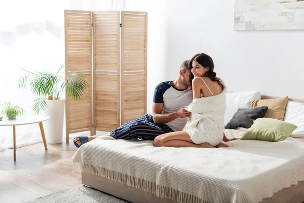 Bearded man kissing smiling girlfriend in cardigan on bed at home - foto de stock