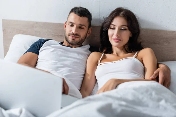 Man hugging girlfriend and looking at blurred laptop on bed — Stock Photo