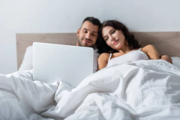 Ordinateur portable près du couple flou couché sur le lit le matin — Photo de stock