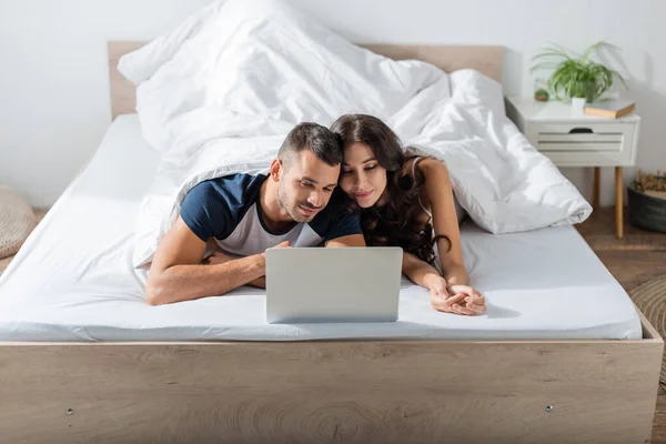 Brunette couple in pajama looking at laptop while relaxing on bed — Stock Photo