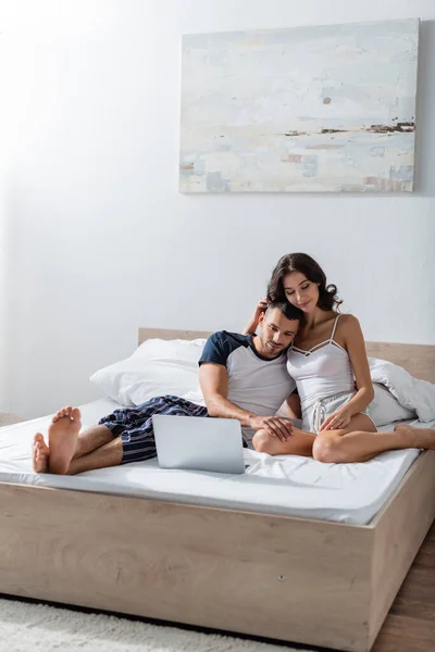 Brunette woman in pajama hugging boyfriend near laptop on bed — Stockfoto