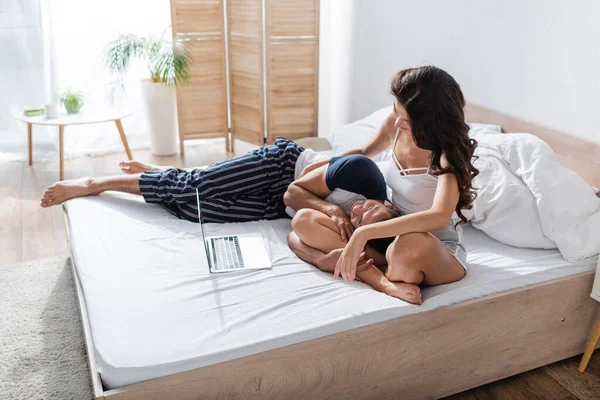 Couple in pajama resting on bed near laptop in bedroom in morning — Fotografia de Stock