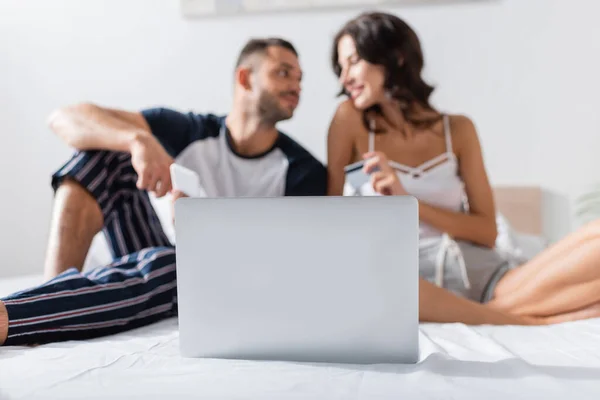 Laptop near blurred couple in pajama sitting on bed — Photo de stock
